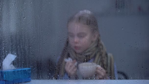 Sick girl sneezing and drinking hot tea behind rainy window, flu virus epidemic — Stock Video