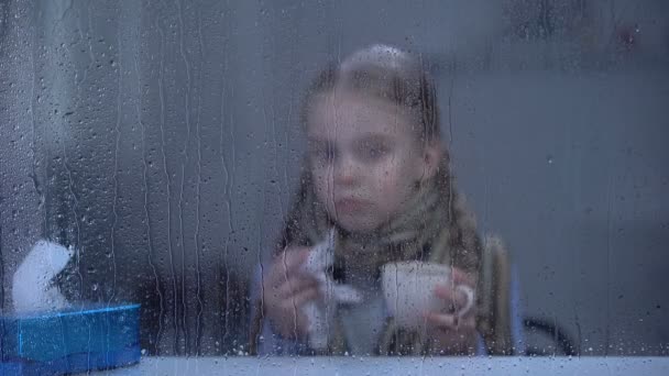 Little girl suffering from flu behind rainy window sneezing and drinking hot tea — Stock Video