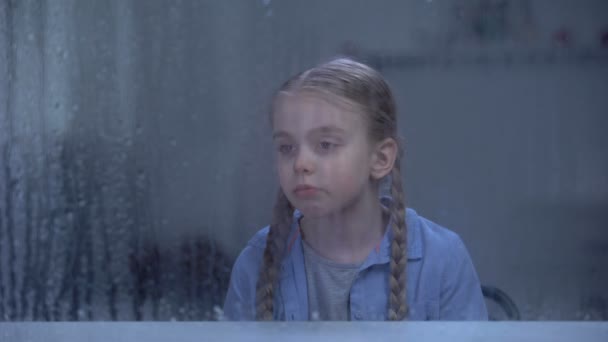 Μοναχικό κορίτσι κοιτάζοντας το παράθυρο σε βροχερό καιρό, περιμένοντας τους γονείς από την εργασία — Αρχείο Βίντεο