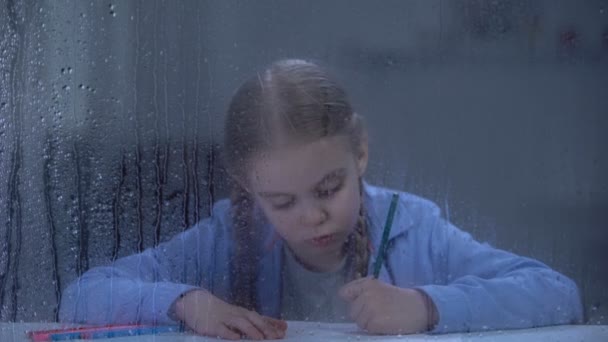 Ωραίο μικρό κορίτσι ζωγραφική πίσω από βροχερό παράθυρο, ορφανό παιδί ονειρεύεται για το σπίτι — Αρχείο Βίντεο