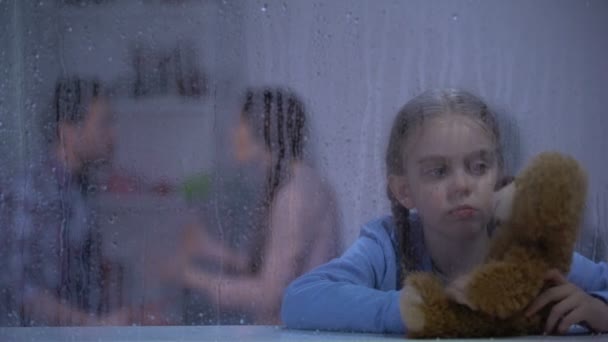 Sad girl with teddy bear on rainy day, father slapping mother on background — Stock Video