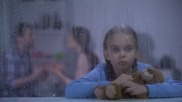 Girl hugging teddy bear on rainy day, father choking mother on background — Stock Video