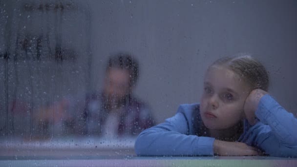 Sad girl sitting behind rainy window, alcohol addicted father drinking in room — Stock Video