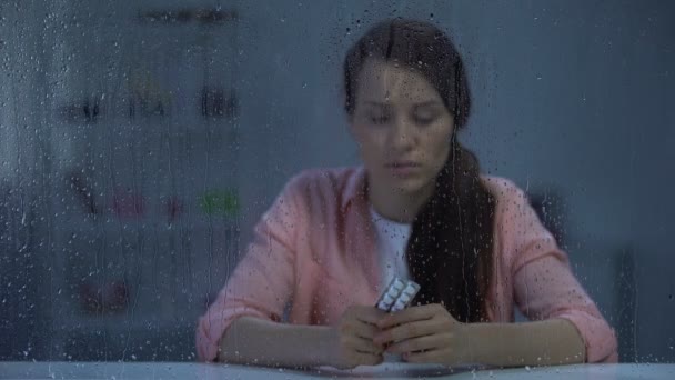 Mujer molesta que sufre de enfermedad fatal sosteniendo píldoras detrás de la ventana lluviosa — Vídeos de Stock