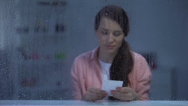 Pensive woman looking at photo behind rainy window, remembering first love — Stock Video