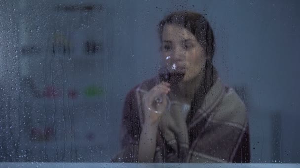 Жінка в ковдрі п'є вино за дощовим вікном, холодний вечір — стокове відео