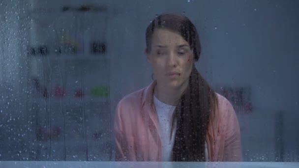 Αναστατωμένη γυναίκα με τραυματισμένο πρόσωπο κοιτάζοντας μέσα από βροχερό παράθυρο, οικογενειακή βία — Αρχείο Βίντεο