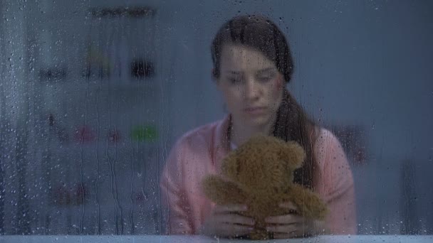 Безнадійна жінка з пораненим обличчям обіймає плюшевого ведмедя за дощовим вікном, підтримка — стокове відео