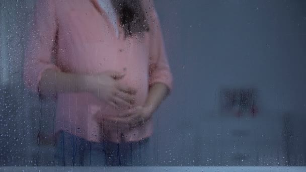 Беременная женщина гладит животик за дождливым окном, ожидает ребенка, материнство — стоковое видео