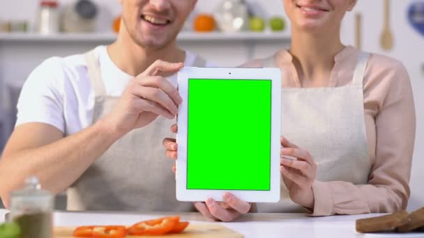 Χαρούμενο ζευγάρι σε ποδιές κρατώντας πράσινη οθόνη tablet κάθεται στην απόδειξη της κουζίνας — Αρχείο Βίντεο