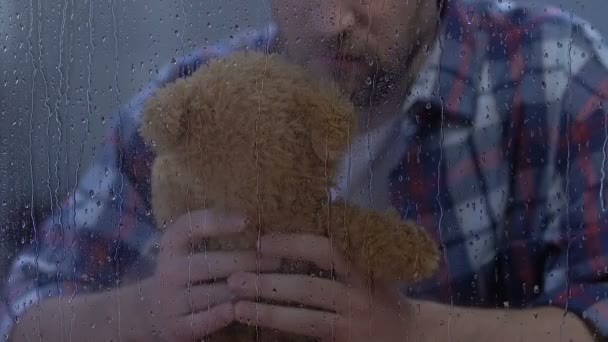 Одинокий мужчина держит плюшевого мишку за дождливым окном, пропавший ребенок после развода — стоковое видео