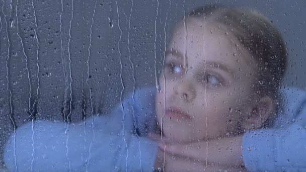 Θλιμμένο κορίτσι που κοιτάζει στο παράθυρο σε σταγόνες βροχής, ονειρεύεται την οικογένεια σε ορφανοτροφείο — Αρχείο Βίντεο