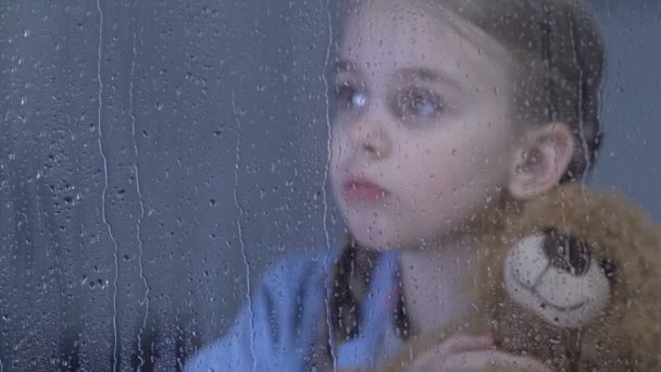 Lonely cute girl hugging teddy bear in orphanage behind rainy window, dreaming — Stock Video