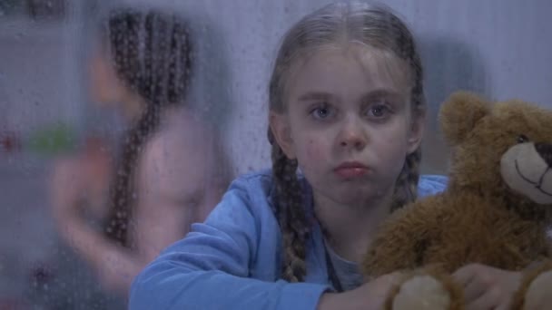 Yağmurlu pencerenin arkasında oturan üzgün kız, kızgın baba anneye saldırıyor, şiddet — Stok video