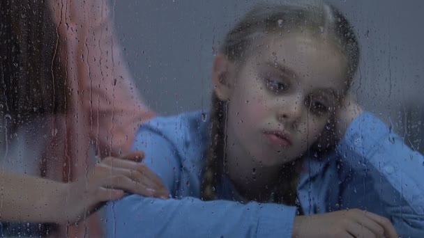 Мати погладжує ніжно засмучену маленьку доньку, що сидить біля дощового вікна, підтримка — стокове відео