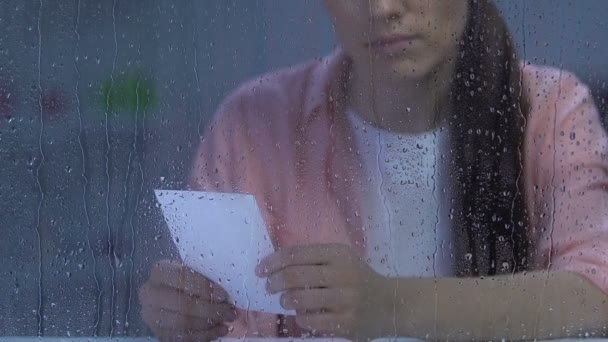 雨の窓の近くで家族の写真を持っている若い女性を動揺させ、離婚に苦しむ — ストック動画