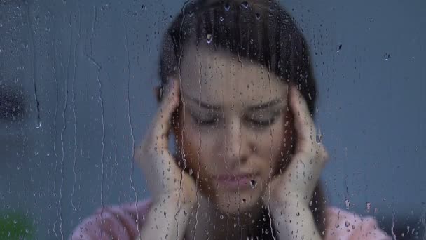 Женщина страдает мигренью, растирает виски возле дождливого окна, чувствительность к погоде — стоковое видео