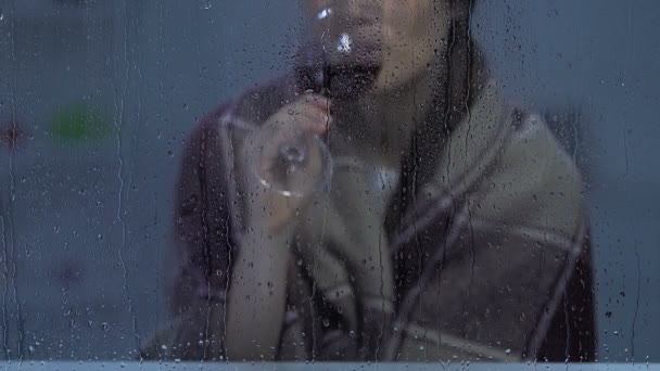 Középkorú nő borított kockás közelében ülve esős ablakon, és iszik bort — Stock videók