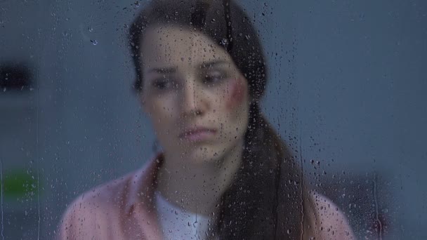 Mujer infeliz tocando mejilla herida detrás de ventana lluviosa víctima de violencia doméstica — Vídeos de Stock