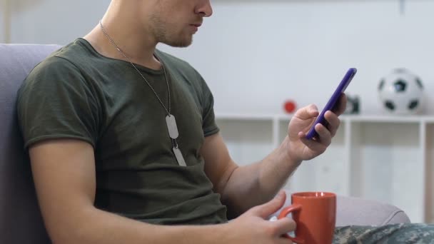 Army sergeant reading smartphone messages holding coffee cup, resting on sofa — Stock Video