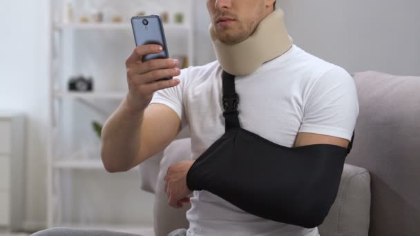 Hombre en cuello cervical y el cabestrillo del brazo chatear smartphone en casa, rehabilitación — Vídeos de Stock