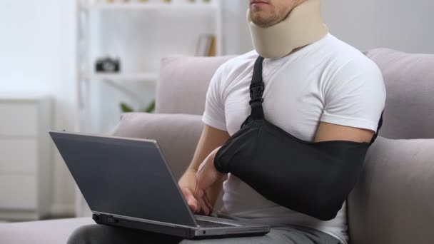 Мужчина в перевязке рук и шейном воротнике, работающий на ноутбуке дома, лечение травм — стоковое видео