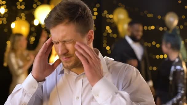 Молодой человек страдает мигренью на вечеринке массируя храмы, раздражая музыку, здоровье — стоковое видео