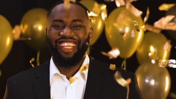 Smiling Afro-American man standing under falling confetti, party decorations — Stock Video