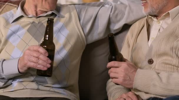 Двое пенсионеров звонят в бутылки, пьют пиво, сидя на диване, свободное время — стоковое видео