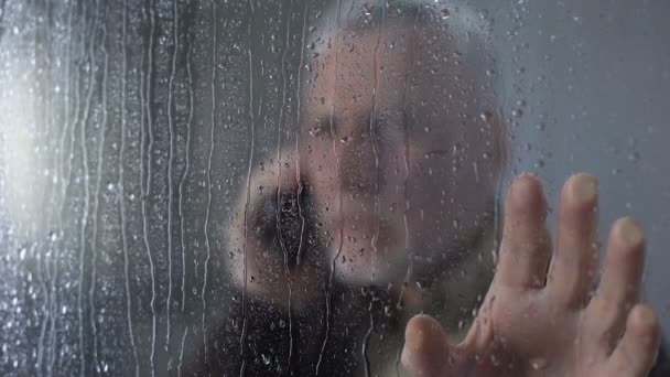 Besorgter älterer Mann schaut durch verregnetes Fenster, telefoniert, wartet — Stockvideo