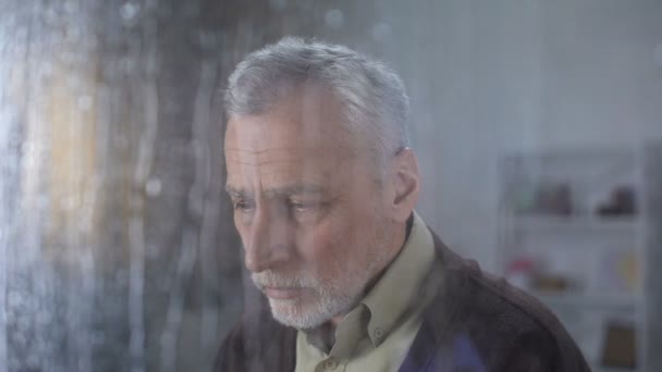 Solitário avô triste olhando na câmera através da janela chuvosa, lar de idosos — Vídeo de Stock