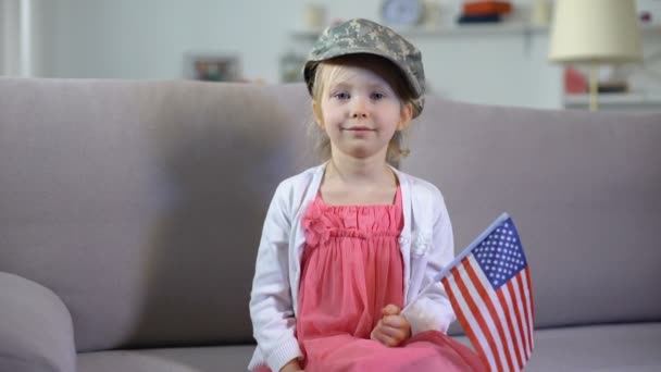 Pequeña patriota con gorra de camuflaje sosteniendo la bandera de Estados Unidos y mirando a la cámara — Vídeos de Stock