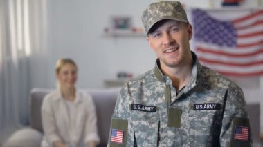 Kamerakarşısında gülümseyen Amerikalı asker, abd bayrağına karşı kanepede oturan gururlu eşi