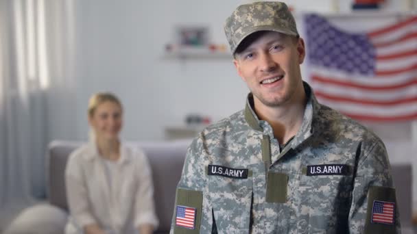 Amerikanischer Soldat lächelt in die Kamera, stolze Frau sitzt auf Sofa gegen uns Flagge — Stockvideo