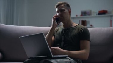 Erkek askeri casus kanepede dizüstü bilgisayar ile oturan ve geceleri akıllı telefon konuşan