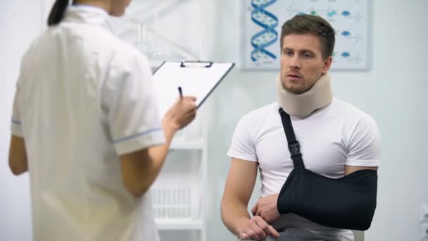 Arzt schreibt Diagnose, um Patientin in Schaumstoff-Halsband und Armschlinge zu verärgern — Stockvideo