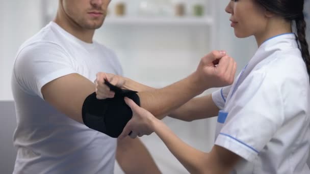 Улыбающийся доктор фиксирует локтевой мягкий ортез порадовал пациента спортсмена, реабилитацию — стоковое видео