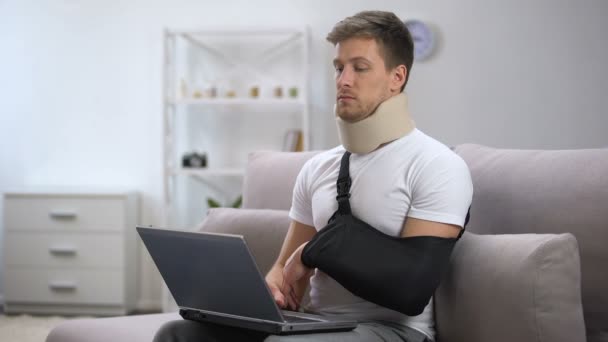 Mannelijke in schuim hals kraag en arm sling typen een hand op laptop, freelance — Stockvideo