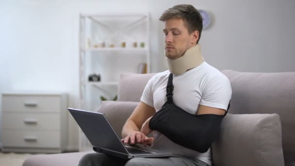 Injured man in foam cervical collar and arm sling working on laptop feeling pain — Stock Video