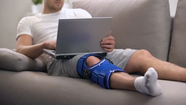 Человек с артритом коленного корсета работает на ноутбуке в период домашней реабилитации и фрилансер — стоковое видео