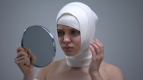 Женщина в эластичной головке, смотрящая в зеркало, чувствует боль после пластической операции — стоковое видео