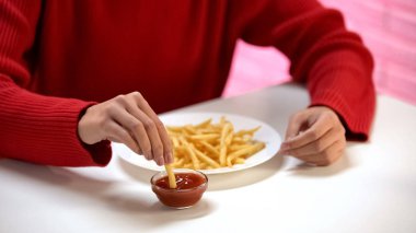 Young lady dipping french-fried potato in tomato sauce, harmful meal, calories