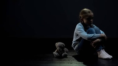 Küçük kız çocuk karanlık odada oyuncak ayı yakınında tek başına oturan, arkadaş eksikliği
