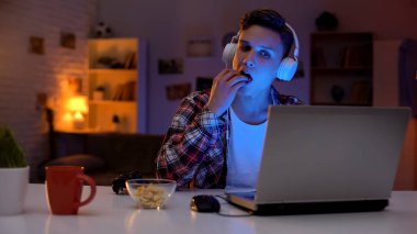 Heyecanlı genç çocuk bilgisayar oyunu oynarken ve atıştırmalık yeme, boş zaman aktivitesi