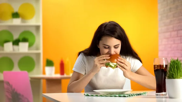 清涼飲料のガラス テーブルの上のカフェでおいしいチーズバーガーを食べて空腹の若い女性 — ストック写真