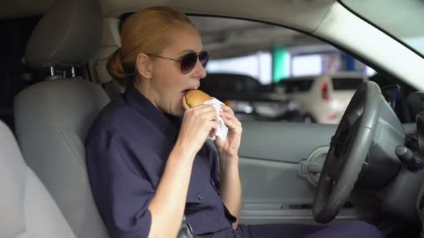 Голодная женщина-коп ест бургер сидя в полицейской машине на парковке, нездоровое питание — стоковое видео
