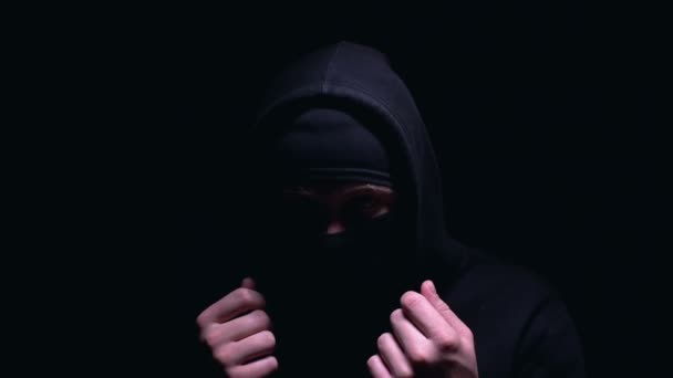 Жуткий человек в балаклаве и капюшоне смотрит в камеру, член бандитской группы — стоковое видео