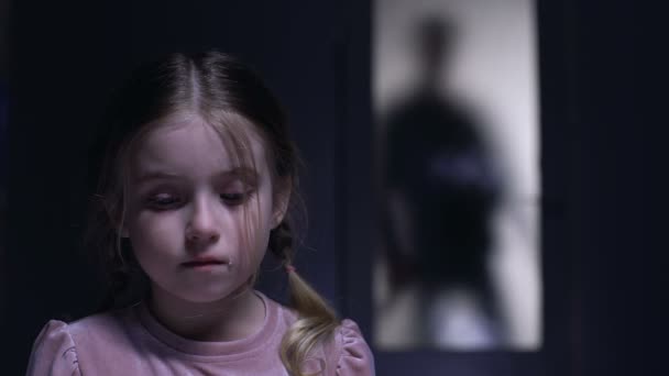 Плачущая маленькая девочка смотрит в камеру силуэт алкоголика отца за дверью — стоковое видео