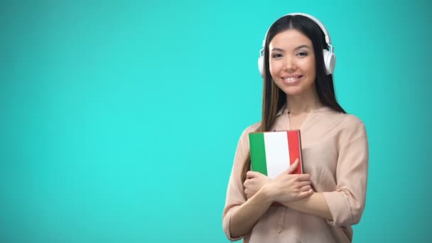 Chica escuchando lecciones de audio italiano en auriculares, libro en las manos, traducciones — Vídeo de stock