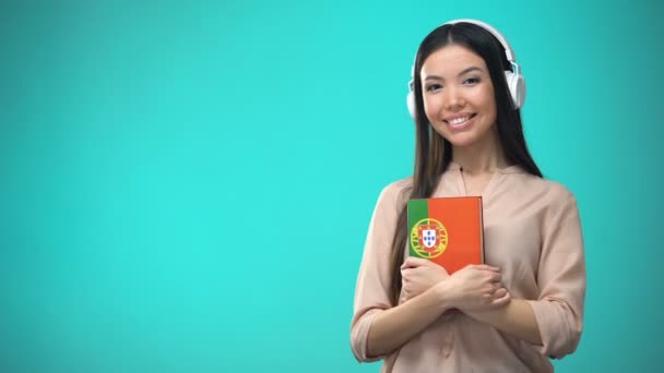 Menina escutando aulas de áudio português em fone de ouvido, livro em mãos, traduções — Vídeo de Stock